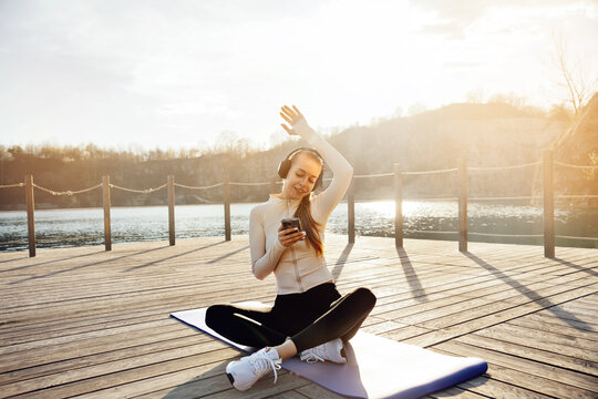 Young woman listening to music during yoga break outdoors. Beautiful woman using smartphone with relaxed smile beside lake. Healthy lifestyle concept.