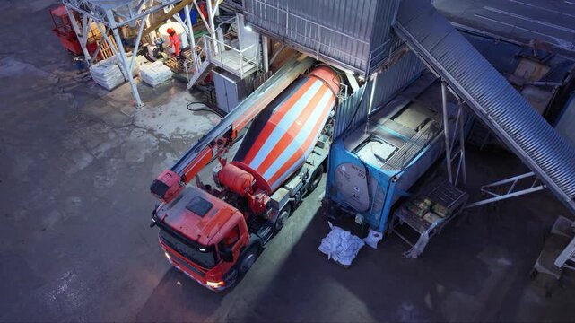 A red concrete mixer truck stands beneath a batching plant loading structure at dusk, with its striped mixing drum turning beside tall silos and illuminated equipment.