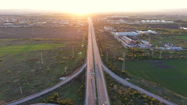 Aerial pullback above a cloverleaf interchange as vehicles follow the curved ramps, with green fields surrounding the roads and the sun low on the horizon.