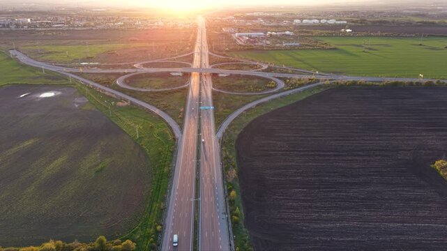 Aerial view of a cloverleaf highway interchange with looping ramps and moving traffic, surrounded by agricultural fields illuminated by low golden evening light.