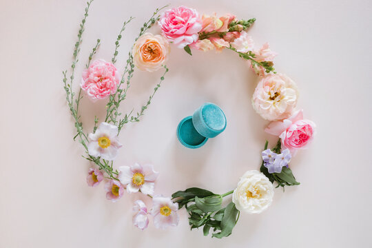 Overhead view of an open teal velvet ring box in the middle of a floral wreath with pink, white, and peach roses and anemones on a white background