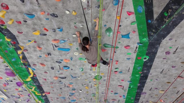 Female rock climber ascending an indoor climbing wall with colorful holds, wearing a safety harness and climbing shoes in a modern climbing gym.
