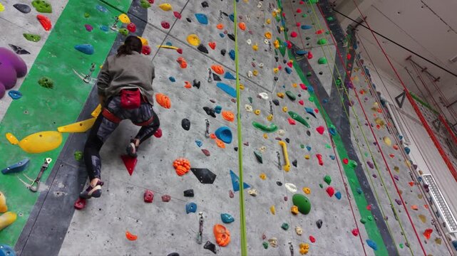 Female rock climber ascending an indoor climbing wall with colorful holds, wearing a safety harness and climbing shoes in a modern climbing gym.