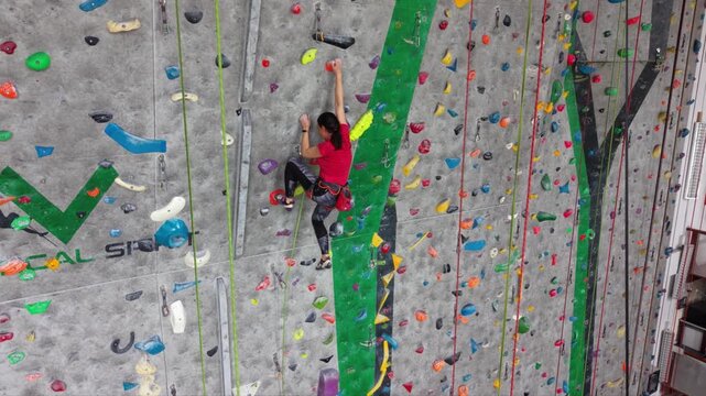 Female rock climber ascending an indoor climbing wall with colorful holds, wearing a safety harness and climbing shoes in a modern climbing gym.
