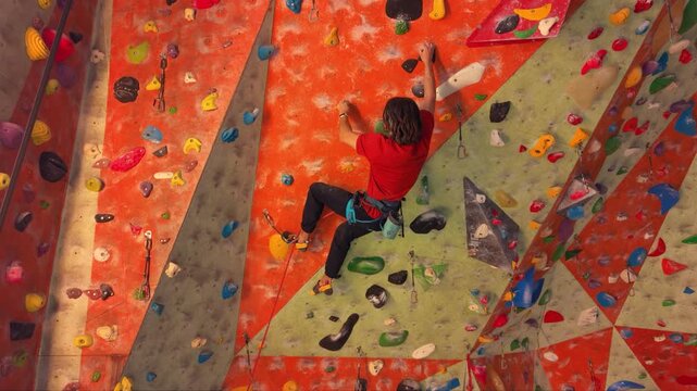 Indoor rock climber ascending a steep artificial climbing wall with colorful holds and volumes while secured by a rope and harness, showing strength, technique and concentration during sport climbing.