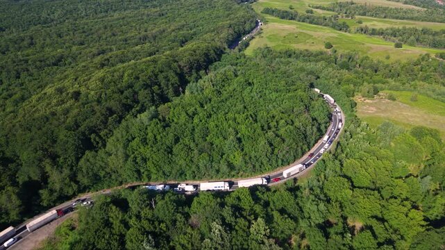 Aerial view of a long traffic queue with trucks and cars moving slowly along a winding two-lane road through dense green forest and rolling countryside, showing heavy congestion on a rural route.