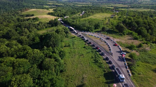 Aerial view of a long traffic queue with cars and heavy trucks on a winding rural mountain road, surrounded by dense green forest and countryside.