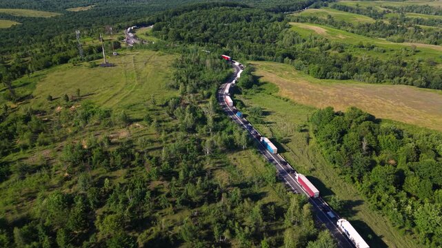 Aerial view of a long traffic queue with cars and heavy trucks on a winding rural mountain road, surrounded by dense green forest and countryside.