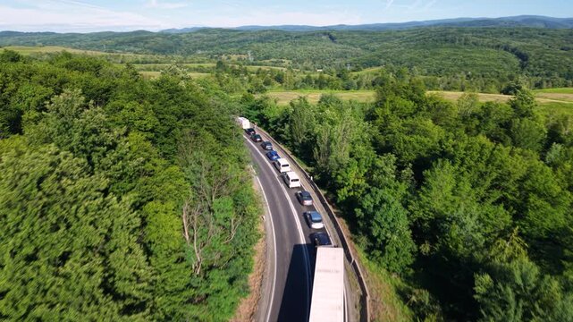Aerial view of a long traffic queue with trucks and cars moving slowly along a winding two-lane road through dense green forest and rolling countryside, showing heavy congestion on a rural route.