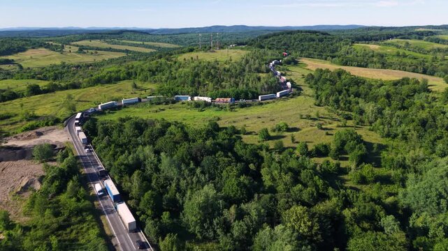 Aerial view of a long traffic queue with trucks and cars moving slowly along a winding two-lane road through dense green forest and rolling countryside, showing heavy congestion on a rural route.