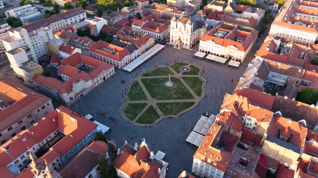 High aerial orbit over a formal city square with patterned green lawns and a central monument, enclosed by historic red roofed buildings and a pale twin tower church.