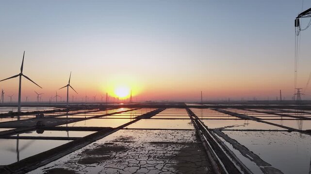Wind turbines aligned over salt pans during sunset with reflective water surfaces and distant horizon