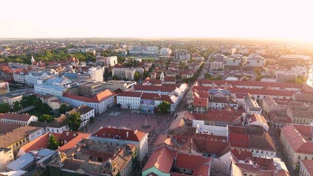 Wide aerial view of red roofed historic blocks surrounding a circular patterned square, with a large theater building and distant urban districts in sunset light.