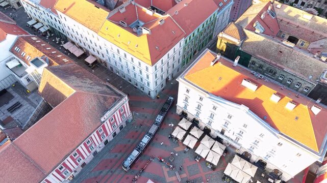 Top-down aerial view of a modern tram travelling through the historic city centre of Timișoara, Romania, crossing a pedestrian square surrounded by traditional European architecture.