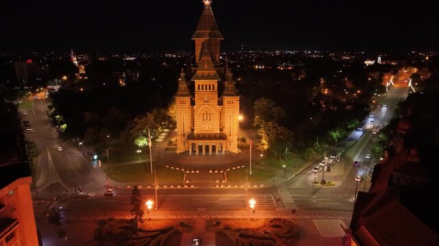 Night aerial view of the illuminated Romanian Orthodox Metropolitan Cathedral in Timișoara, Romania, surrounded by city streets, traffic and urban lights in the historic city centre.