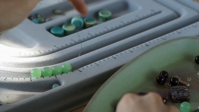 Hands thread green beads onto a needle. Beads rest on a gray organizer tray to make jewelry, specifically beading. 