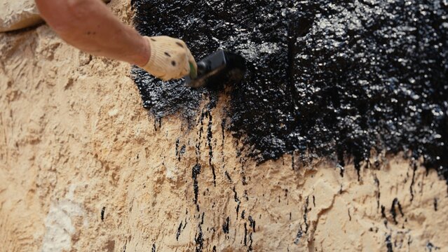 Construction worker in protective glove applying black liquid bitumen mastic to a rough concrete foundation wall, providing essential waterproofing and protective coating for the building structure