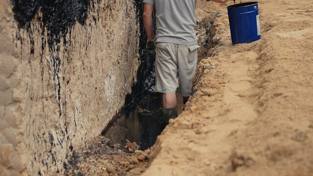 Worker standing in a trench, carefully applying a black bituminous waterproofing compound to the concrete foundation wall with an applicator, ensuring a protective barrier against moisture