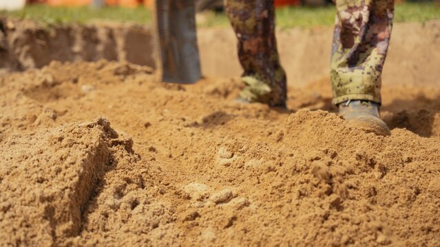 Person wearing camouflage pants and work boots standing in a trench, using a shovel to dig and move earth, creating a mound of sand for excavation work