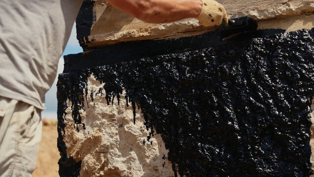 Construction worker applying a liquid bituminous coating with a brush to a stone wall foundation, creating a protective barrier against moisture and water damage