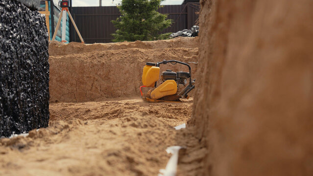 Yellow plate compactor resting on sand, preparing the ground for a new building's foundation within an excavated pit at a construction site