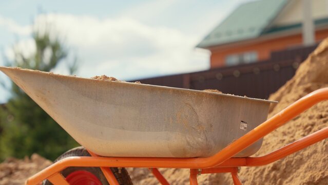 Orange wheelbarrow carrying a pile of sand, prepared for building work or home renovation project, with a house roof in the blurred background