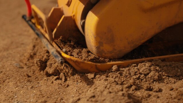 Plate compactor vibrating and compressing loose dirt and sand on a construction site, preparing the ground for foundations, paving, or landscaping with heavy machinery