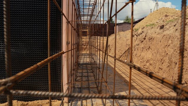 Steel rebar cage forming the reinforcement for a building foundation, prepared for concrete pouring in a construction trench with excavated soil and a waterproof wall