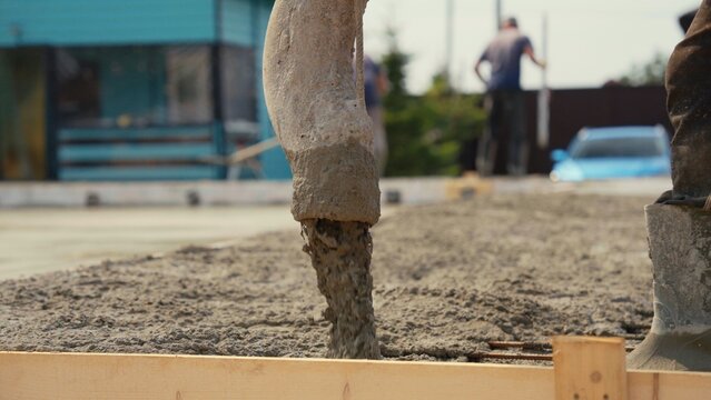 Concrete pump hose pouring fresh cement mix into a prepared wooden formwork for a building foundation, with workers in the background