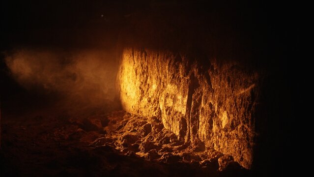 Dark cavern showing a rough earth wall illuminated by a golden light source, with smoke wafting through the deep shadows, creating an atmospheric and textured subterranean scene