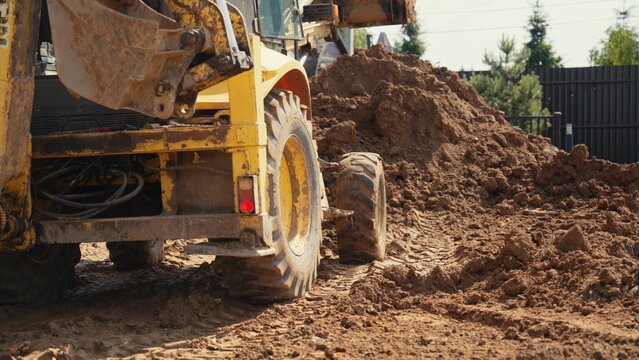 Yellow construction machinery performing earthworks, moving large piles of dirt and soil, creating tire tracks on the ground during a sunny day
