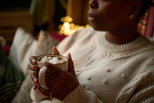 African American young adult woman holding mug of hot chocolate with marshmallows, relaxing at home. Image represents winter comfort, self-care, and holiday season marketing, close up