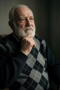 Elderly man with white beard rests chin on hand. He wears argyle sweater, looks away thoughtfully. Man sits indoors, subdued light creates deep shadows and quiet mood.