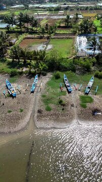 Aerial view of traditional fishing boats moored on the muddy shore with lush agricultural fields and palm trees in the background in Kecamatan Kebumen, Jawa Tengah, Indonesia.