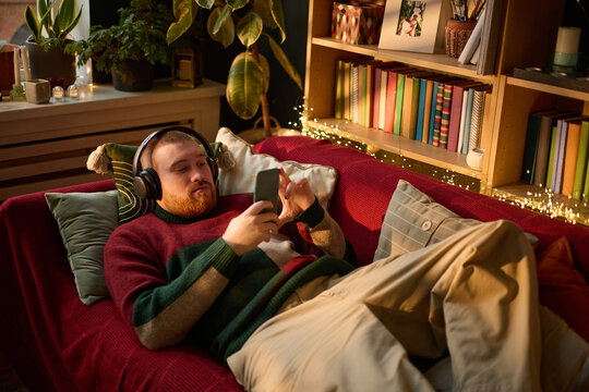 Caucasian young adult man lying on sofa wearing headphones and using smartphone. Image represents digital entertainment and leisure, suitable for mobile app marketing campaigns