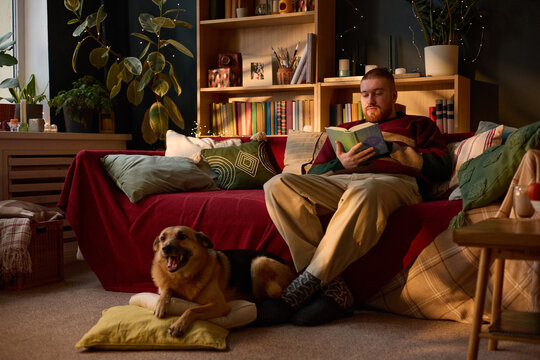 Young adult Caucasian man sitting on sofa and reading book with dog lying on floor nearby, representing leisure and mental wellbeing. Image suitable for pet care and lifestyle marketing