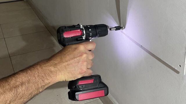 Close-up of construction worker's hand using a cordless screwdriver to fasten a screw into drywall, led light illuminating the mounting area during a home renovation project.