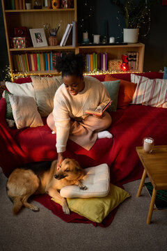 Vertical image of young adult African American woman sitting on sofa, reading book and petting her dog. Scene represents pet ownership and is ideal for lifestyle marketing
