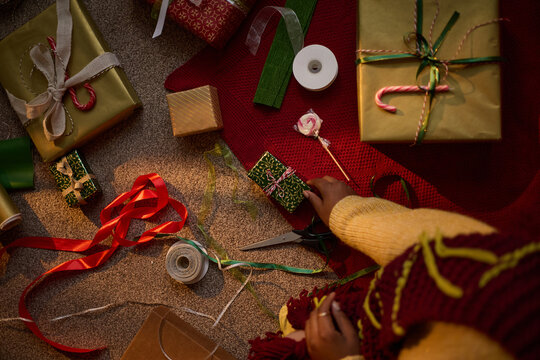 African American woman wrapping Christmas gifts on floor, preparing holiday presents with ribbons and scissors. Image ideal for festive retail marketing and seasonal greeting campaigns