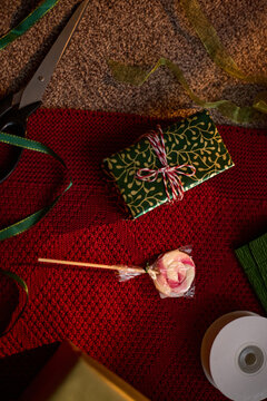 Wrapped gift box, lollipop, and scissors lying on red knitted blanket, representing holiday preparation. Ideal for Christmas marketing campaigns, vertical