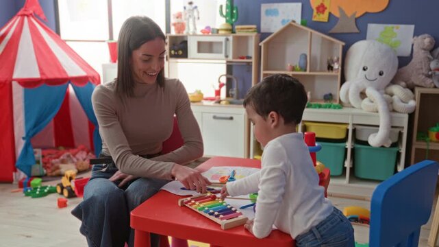 Woman smiles as little boy hands a xylophone mallet across a red table while reaching with his hand in a preschool classroom; playful learning joy.