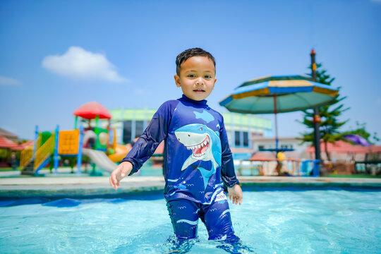 A cheerful Asian boy in a blue shark swimsuit smiles while playing in a sunny outdoor swimming pool at a resort.