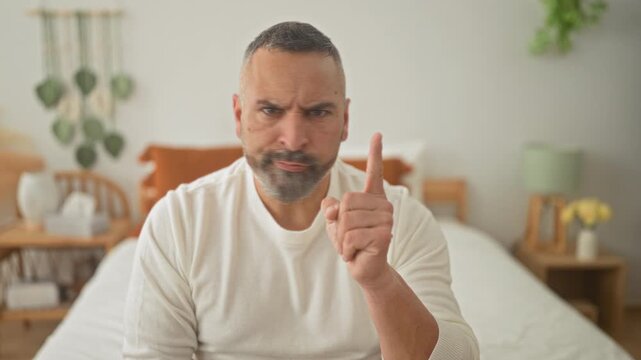 Man points index finger up while seated on bed in a bedroom, bearded, scowling and gesturing for emphasis; stern warning.