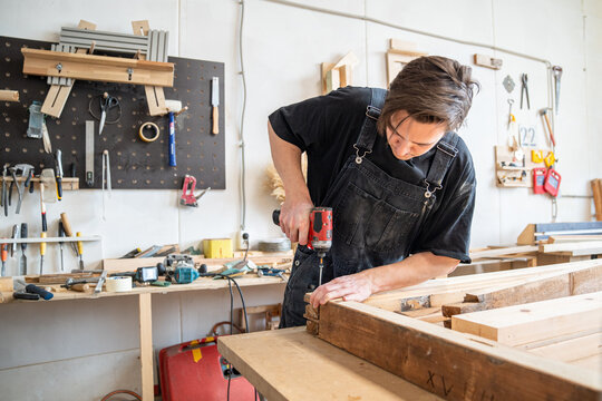 Woodworker technician using handheld cordless electric driver to fasten long screws in thick wooden frame, carpenter completing assembling stage and fastens frame parts together with drill tool.