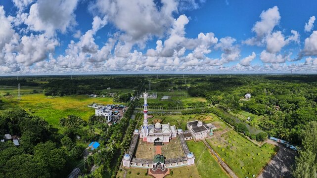 Aerial view of the Baitul Aman Jame Masjid Complex surrounded by lush green gardens, agricultural fields, and trees under a bright blue sky in Barishal, Barisal Division, Bangladesh.