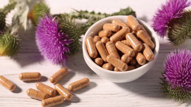 Milk thistle pills and flowers on white wooden table, closeup. Camera moving right