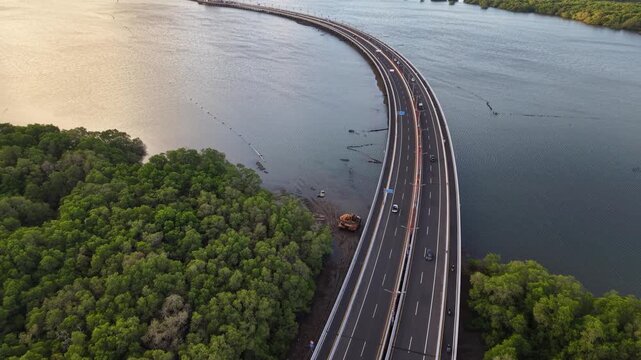 Aerial view of the Bali Mandara Toll Road curving through lush mangrove forests over the water in Kabupaten Badung, Bali, Indonesia.