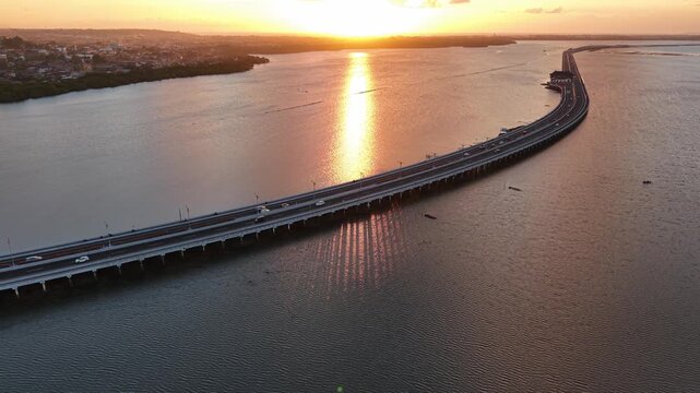 Aerial view of Mandara Toll Road stretching across the water at sunset with golden light reflecting on the surface in Kabupaten Badung, Bali, Indonesia.