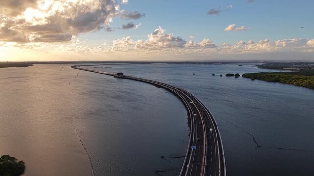 Aerial view of the Mandara Toll Road stretching across the water at sunset with golden light reflecting on the surface and mangrove forests in Kabupaten Badung, Bali, Indonesia.