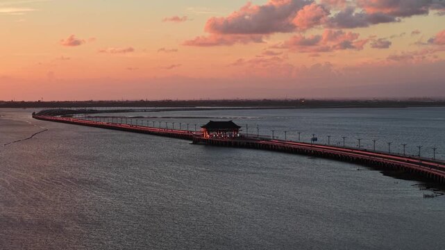 Aerial view of the Bali Mandara Toll Road stretching across the ocean at sunset with warm light reflecting on the water in Kabupaten Badung, Bali, Indonesia.